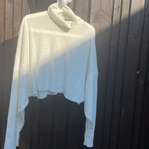 Vit croppad polotröja med ribbad struktur - Säljer en vit croppad polotröja med ribbad struktur och långa ärmar. Tröjan har en lös passform och hög polokrage, perfekt för lager-på-lager och chill vibes. Asymmetrisk nederkant ger en cool och avslappnad look.