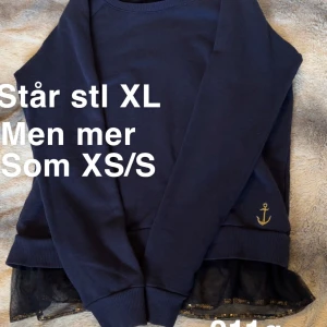 Mörkblå tröja med ankardetalj - Mörkblå tröja med rund hals och långa ärmar. Nedtill finns en svart tyllvolang med små guldiga paljetter och en broderad guldfärgad ankare på ena sidan. Perfekt för dig som gillar marina detaljer och en unik touch.🌸Står stl XL men väldigt liten. Mer som XS/S