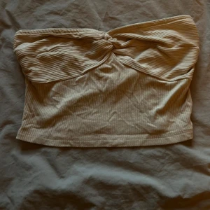 Beige ribbad bandeau tubtopp - Säljer en beige bandeau tubtopp med ribbad struktur och snygg twistdetalj framtill. Toppen är axelbandslös och har en croppad passform som passar perfekt till höga jeans eller kjol.
