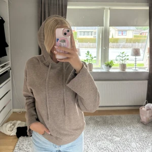 Beige stickad hoodie med huva - Mysig beige stickad hoodie med huva och snörning. Tröjan har en relaxed passform och långa ärmar, perfekt för kyliga dagar. Passar lika bra till jeans som till mjukisbyxor. Enkel och stilren design som funkar till det mesta.