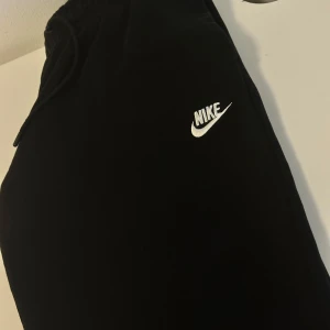 Svarta Nike mjukis  - Svarta mjukis från Nike med klassisk vit logga broderad på ena benet. Mjuk och skön bomullskänsla, elastisk midja med snörning för perfekt passform. Perfekta för chill eller träning, enkel och stilren design.