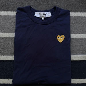 Mörkblå t-shirt Comme des Garçons Play - Snygg mörkblå t-shirt från Comme des Garçons Play med det ikoniska gula hjärtat broderat på bröstet. Klassisk rund halsringning och korta ärmar. Perfekt för dig som gillar stilrena plagg med en twist.