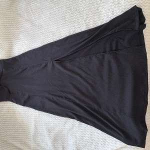 Svart långklänning med volangärm - Supersnygg svart långklänning från lager 157 med volangärmar och söt knytning framtill. Klänningen har en mjuk, lätt ribbad struktur och en feminin passform som faller snyggt. Perfekt för dig som gillar en stilren look med en twist.