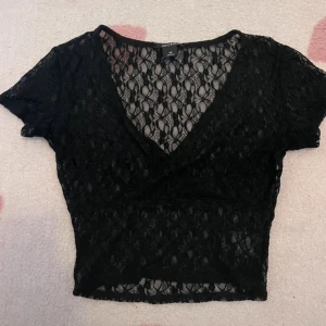 Svart spetsig croppad omlottopp - Svart croppad topp från Monki i  spets med omlott och v-ringad hals. Bara använt ett par fåtal gånger. Som ny. 