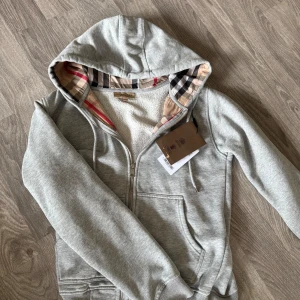 Burberry hoodie - Grå hoodiejacka från Burberry med dragkedja och klassiskt rutigt foder i beige, svart och rött. Jackan har huva, mjukt fleecefoder och ribbade muddar. Perfekt för dig som vill ha en stilren och bekväm look med lyxig känsla.