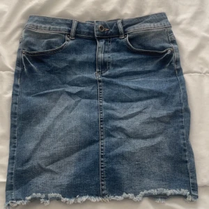 Jeanskjol från Pieces med fransig kant - Snygg blå jeanskjol från Pieces i klassisk femficksmodell. Kjolen har fransig nederkant, bälteshällor och knappstängning framtill. Perfekt för dig som gillar en avslappnad och trendig stil. Modellen är rak och slutar ovanför knäna.