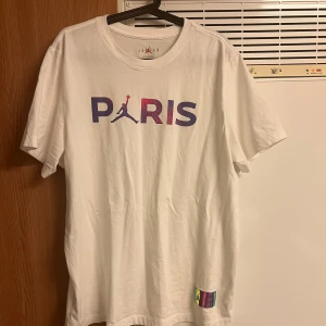 Vit PSG x Jordan t-shirt - Vit t-shirt från Jordan och PSG med coolt Paris-tryck i lila och rosa på bröstet och stor logga på ryggen. Holografisk patch nertill med Jordan och PSG-logga. Klassisk passform och rund hals, perfekt för dig som gillar streetwear och sport.                        Det är nästan ny å använd bara2-3 gånger (St L men passar till de som har XL i storlek.  750kr per ink frakt 