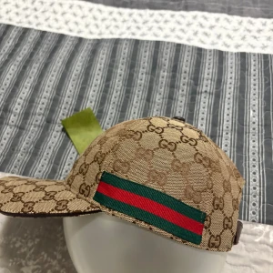 Gucci keps med GG-mönster och röd/grön detalj - Exklusiv keps från Gucci i beige och brunt med klassiskt GG-monogram över hela kepsen. Snygg röd och grön rand på sidan och justerbart brunt läderband bak. Perfekt för dig som vill sticka ut med en lyxig accessoar.