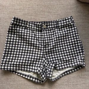 Svartvita rutiga shorts från H&M Divided - Rutiga shorts från H&M Divided i svart och vitt. Klassisk modell med korta ben,  hög midja, bälteshällor, knapp och dragkedja framtill samt två bakfickor. Gjorda i mjuk bomull med lite stretch för extra komfort. Perfekta för en chill sommardag. Stl 40