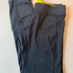 Svarta träningsleggings från Newbody - Säljer ett par svarta träningsleggings från Newbody. De är tillverkade i ett stretchigt material med 92% polyamid och 8% elastan. Perfekta för träning med en bekväm passform och elastisk lågmidja.  Gul inuti linning  Liten ficka framtill i linning Använda och tvättade 3 gånger  Inga skador eller anmärkningar ( kan finnas några små noppror men syns inte)  Byxans totala längd 85 cm  Innerbenslängd 70 Resårkanten är cirka 5 cm bred    Mycket fint skick 