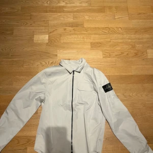 Vit overshirt från Stone Island - Snygg vit overshirt från Stone Island med klassisk krage, bröstficka och svart dragkedja framtill. Ikonisk logga på vänster ärm. Perfekt för lager-på-lager och streetwear-vibe.