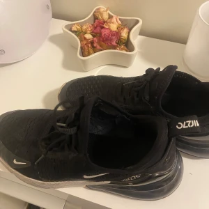 Svarta Nike Air Max 270 sneakers - Svarta Nike Air Max 270 sneakers med stora luftbubblor i sulan och vita detaljer. Ovandelen är i mesh för bra andningsförmåga och skorna har snörning. Perfekta för dig som gillar sportig och modern stil.