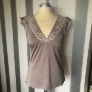 Beige  topp med spets från Saint Tropez - Supersöt beige topp  från Saint Tropez med v-ringning och dekorativ spetskant i vitt. Ärmlös modell med små volangdetaljer på axlarna. Blusen har en mjuk, glansig yta och känns riktigt feminin och romantisk.