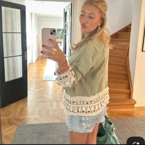 Grön jacka med virkade detaljer - Säljer en ljusgrön overshirt med stora knappar och unika virkade detaljer med tofsar och pärlor längs ärmslut och nederkant. Jackan har en loose passform och är perfekt för dig som vill sticka ut med en boho-inspirerad look.