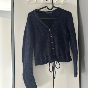 Mörkblå kofta från Brandy Melville - Supermysig mörkblå stickad kofta från Brandy Melville med v-ringning, knappar framtill och snörning i midjan. Långärmad och lite croppad modell som är perfekt att slänga över en topp. Materialet är mjukt och fluffigt, perfekt för kyligare dagar.