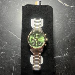 Dam kronografklocka BOSS - Stilren klocka från BOSS med grön urtavla, guldiga detaljer och silverfärgad länk i ädelstål. Klockan är både vattentät samt har ett viklås. Klockan har tre subdials, datumvisning och kronograf-funktion. Perfekt accessoar för dig som vill sticka ut med en snygg och modern look.