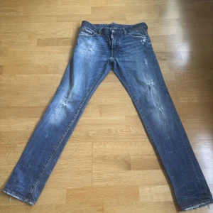 Blå Dsquared2 jeans, storlek 46 - Säljer ett par blå Dsquared2 jeans i storlek 46. Jeansen har slitna detaljer och en snygg tvätt med ljusare partier. Klassisk femficksmodell med Dsquared2-logga vid gylfen och silverfärgad knapp. Tillverkade i Italien i mjukt bomullsjeansmaterial.