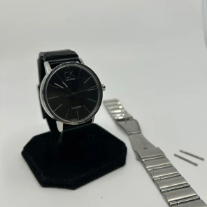 Calvin Klein klocka med stållänk - Stilren klocka från Calvin Klein med svart urtavla och silverfärgade detaljer. Klockan har ett svart läderarmband och medföljer även en utbytbar länk i silverfärgat stål. Klassisk rund form och minimalistisk design, perfekt för dig som gillar enkelhet och elegans.
