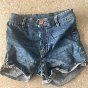 Blå jeansshorts med upprullad kant - Klassiska blå jeansshorts med upprullad kant och femficksmodell. De har bälteshällor, knapp och dragkedja framtill samt två bakfickor. Perfekta för varma dagar och enkel att matcha med allt.