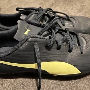 Svarta fotbollsskor från Puma - Snygga svarta fotbollsskor från Puma med gula detaljer och klassisk logga på sidan och plös. Skorna har snörning och mönstrad ovandel i syntetmaterial. Yttersulan har fasta dobbar i gult och svart för bra grepp på planen.