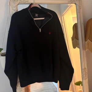 Svart merinoullströja med half zip - Svart långärmad tröja i mjuk merinoull med half zip och liten röd logga på bröstet.  