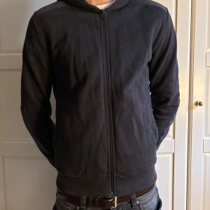 Svart Colmar cardigan med huva - Svart bomberjacka från Colmar med huva och hel dragkedja framtill. Jackan har en stilren look, ribbade muddar och en diskret logga på ärmen. Perfekt för dig som gillar enkel och clean stil. Modellen är 193, men tröjan är lite för liten.