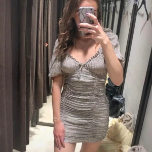 klänning Zara - Supersöt kort klänning från Zara i beige med svarta prickar. Klänningen har puffärmar, v-ringning med liten knytning framtill och smockad passform som sitter tajt. Perfekt för dig som gillar trendiga och feminina detaljer!