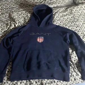 Marinblå hoodie från GANT - Snygg marinblå hoodie från GANT med broderad logga och sköld på bröstet. Storlek 170 cm och 15 år. Inga fläckar och inga slitna ställen. Kan gå lägre i pris.