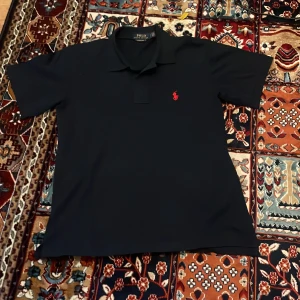 Mörkblå Polo Ralph Lauren pikétröja - Mörkblå pikétröja från Polo Ralph Lauren i custom slim fit. Klassisk krage, korta ärmar och röd broderad logga på bröstet. Tillverkad i bomull för en skön känsla och snygg passform. Perfekt för en clean och stilren look.