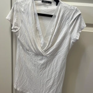 Vit omlottopp från Esprit - Vit omlottopp från Esprit i mjuk bomull. T-shirttyp med kort ärm och djup v-ringning framtill som ger en snygg draperad effekt. Perfekt att styla med jeans eller kjol för en clean look.