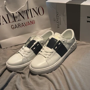 Valentino Garavani Open sneakers vit/blå - Snygga Valentino Garavani Open sneakers i vitt skinn med bred marinblå rem över mitten. Klassisk rund tå, vita snören och ikoniska nitar på hälen. Lyxig känsla och stilren design som sticker ut.