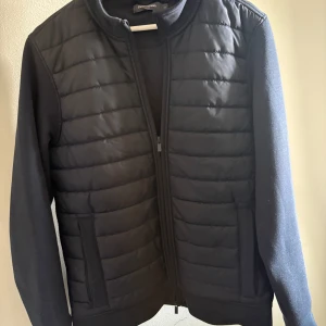 Mörkblå cardigan  - Mörkblå pufferjacka/cardigan från Dressman med quiltad framsida och släta ärmar. Jackan har dragkedja framtill, ståkrage och två fickor. Perfekt för dig som gillar stilren och enkel design.