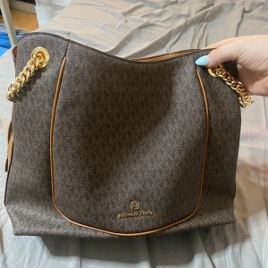 Brun axelväska Michael Kors - Snygg brun axelväska från Michael Kors med MK-logga över hela väskan. Väskan har guldfärgade kedjedetaljer, bruna skinnremmar och flera fack inuti, inklusive dragkedjefack. Perfekt storlek för allt du behöver och stilren design. Den är äkta jag har tappat bort kvittot , den är köpt på Michael kors butiken ! Jag har använt den 3 gånger !