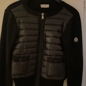 Svart Moncler cardigan jacka - Svart cardigan jacka från Moncler med quiltad front i blankt material och stickade ärmar. Två fickor framtill med knappar och dragkedja hela vägen upp. Klassisk Moncler-logga på ärmen. Perfekt för dig som vill ha en stilren och exklusiv look.