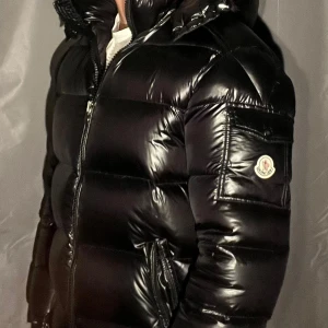 Svart Moncler pufferjacka med huva - Säljer en svart, glansig pufferjacka från Moncler med huva och dragkedja framtill. Jackan har Moncler-logga på ärmen, praktiska fickor och en riktigt fet, vadderad look som håller dig varm. Perfekt för dig som vill ha en stilren och exklusiv vinterjacka. Kommer med några defekter på framfickan och ryggen (se dom sista bilderna) där av det billiga priset