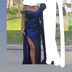 Marinblå långklänning  - Elegant marinblå långklänning från Sasha med figurnära passform och draperad detalj framtill. Klänningen har breda axelband och en snygg byst med markerade kupor. Tillverkad i glansigt satinliknande material som ger en lyxig känsla.