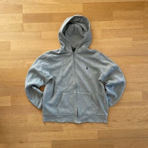Grå zip up hoodie från Polo Ralph Lauren - Klassisk grå hoodie från Polo Ralph Lauren med dragkedja framtill och broderad marinblå logga på bröstet. Tröjan har huva och är tillverkad i mjuk bomullsmix. 