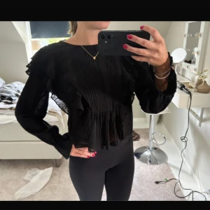 Svart volangblus med transparenta detaljer - Supersnygg svart blus med volanger och transparenta partier. Blusen har lång ärm, lätt rynkade detaljer och en luftig känsla. Perfekt för dig som gillar en trendig och feminin look med extra volym på axlarna.