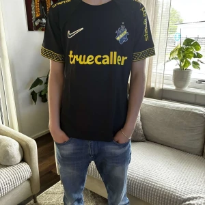 AIK svart matchtröja Craft 2024 - AIK:s officiella svarta matchtröja från 2024 med gula detaljer och tryck från Truecaller, Stadium och SVEA. Klassisk AIK-logga på bröstet, korta ärmar med unikt gult mönster vid ärmslut. Tillverkad i lätt och ventilerande funktionsmaterial.