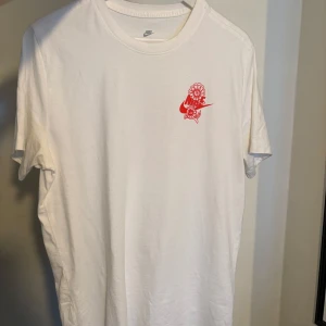 Vit Nike t-shirt chinese new year - Vit t-shirt från Nike med röd grafisk print framtill och stor ryggdesign med tempel och Nike-logga. Klassisk passform och rund halsringning. Perfekt för dig som gillar streetwear och snygga detaljer.