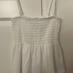 Vit smockad linnetopp med volang - Supersöt vit linnetopp med smockad överdel och volang nertill. Justerbara axelband som går att ta av för att göra toppen axelbandslös. Från gina tricot