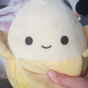 En squismallow-banan - Bilderna visar en mjuk Squishmallows-gosedjur, troligen en bananfigur, med ett glatt ansikte. Ingen bok syns på bilderna.