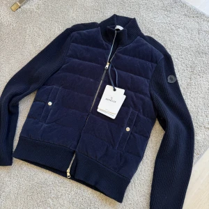 Mörkblå cardigan jacka från Moncler - Snygg mörkblå cardigan jacka från Moncler med quiltad front i manchester och stickade ärmar. Jackan har dragkedja, två fickor med tryckknappar och Moncler-logga på ärmen. 