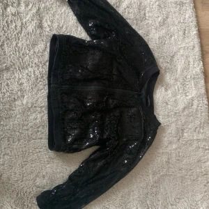Svart paljettjacka från KappAhl - Snygg svart jacka från KappAhl med glittriga paljetter över hela ytan. Jackan har dragkedja framtill och är långärmad med en croppad passform. Perfekt för dig som vill sticka ut och addera lite extra bling till din outfit.