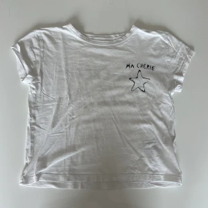 Vit t-shirt med tryck Ma Chérie - Vit t-shirt i bomull med svart tryck fram och bak där det står 'MA CHÉRIE' och en stjärna. Modellen är kortärmad med klassisk rund hals och avslappnad passform. Tröjan är använd men som ny.