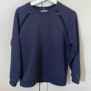 Mörkblå sweatshirt Tommy Hilfiger S - Mörkblå sweatshirt från Tommy Hilfiger med coola håldetaljer längs axlarna. Tröjan har rund hals, långa ärmar och liten logga på ärmslutet. Tillverkad i mjuk bomull och polyester, perfekt för en chill och stilren look.