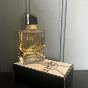 Yves Saint Laurent Libre EdP 90ml - YSL Libre Eau de Parfum i lyxig, genomskinlig glasflaska med guldiga detaljer och svart asymmetrisk kork. Flaskan pryds av den ikoniska YSL-loggan i metall. Doften är elegant och modern, perfekt för dig som gillar exklusiva parfymer.