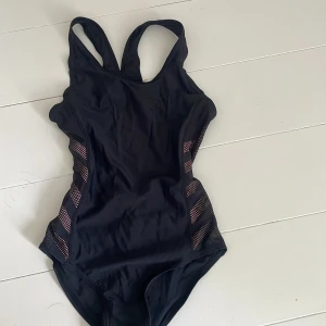 Svart baddräkt med rosa detaljer - Sportig svart baddräkt med breda axelband och öppen rygg. Sidorna har inslag av rosa prickiga meshpaneler i diagonala ränder som ger en cool kontrast. Perfekt för simträning eller strandhäng. Materialet är stretchigt och snabbtorkande syntet. 