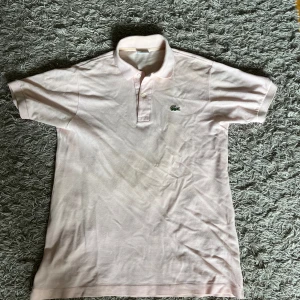 Lacoste piké - Snygg ljusrosa pikétröja från Lacoste med klassisk krage och knäppning framtill. Ikoniska gröna krokodilloggan broderad på bröstet. Tillverkad i bomull med korta ärmar och relaxed fit. Perfekt för en clean och sportig look.