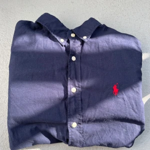 Mörkblå Linneskjorta från Polo Ralph Lauren - Klassisk mörkblå skjorta från Polo Ralph Lauren med röd broderad logga på bröstet. Skjortan har lång ärm, knappar framtill och är tillverkad i mjuk bomull. Perfekt för dig som gillar stilrena skjortor